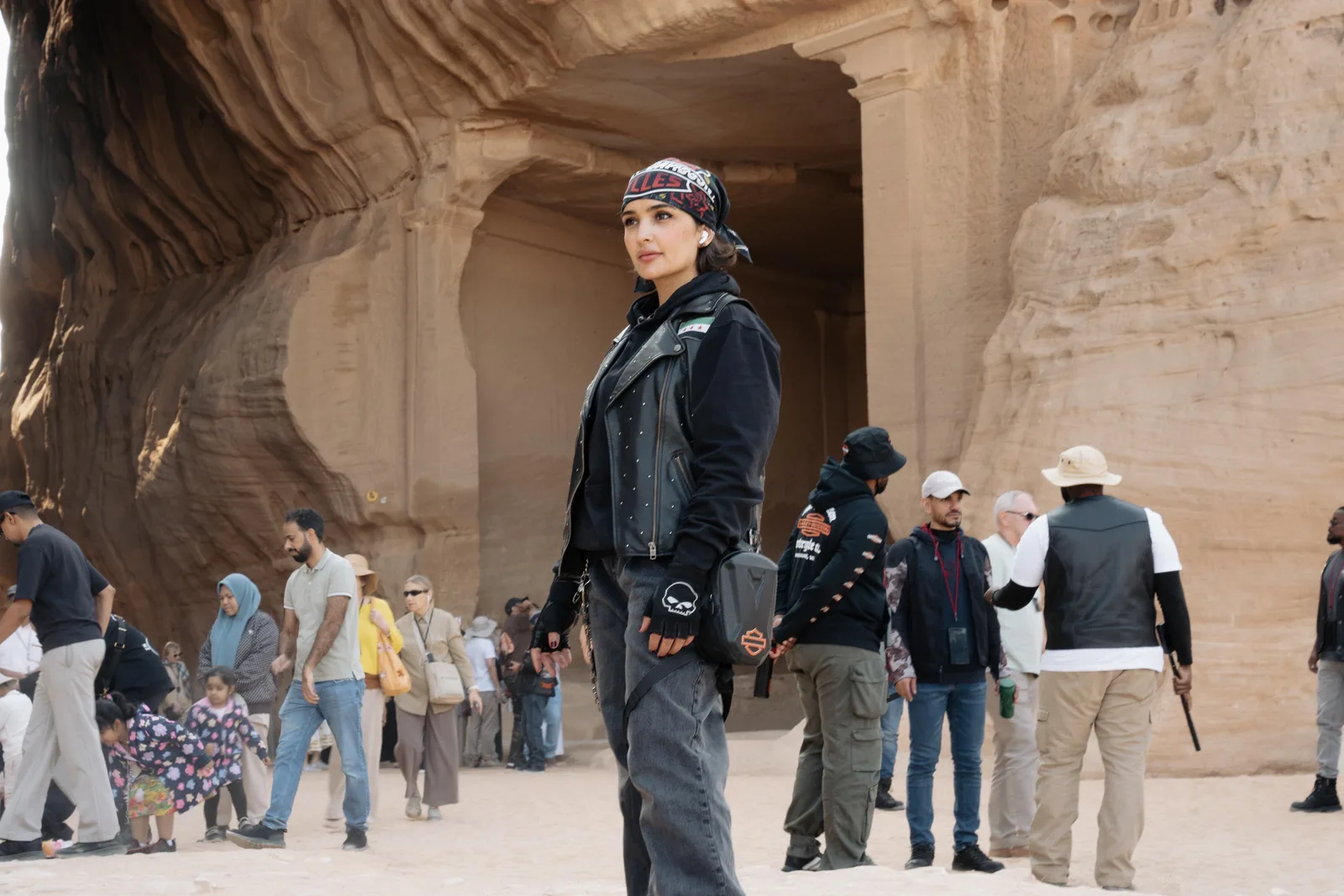 A woman in a black leather vest and bandana stands before a wide chamber carved into a rock face, with visitors of all ages, children among them, behind her.